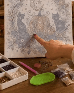 A close-up of a diamond painting or canvas art kit with a hand pointing at a detailed cat and floral design. The scene includes a tray of beads, a pink-handled tool, a green palette, and loose beads on a wooden surface.
